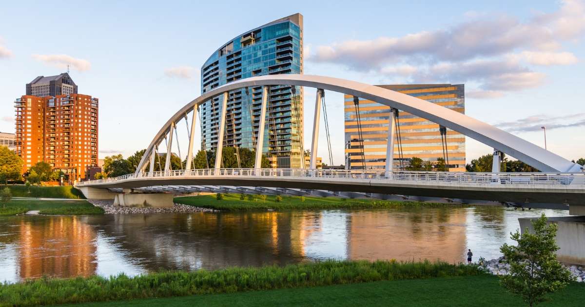 A view of the distinctive white, single-arch Main Street Bridge spanning the Scioto River in downtown Columbus, with modern high-rise buildings and luxury apartments in the background under a partly cloudy sky.