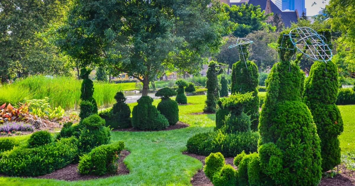 A lush green landscape in The Topiary Park featuring meticulously sculpted evergreen shrubs shaped like people standing, sitting, and strolling through the garden, recreating a famous post-Impressionist painting.