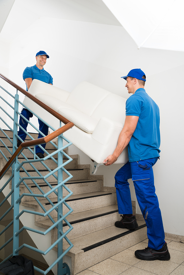 Two Happy Male Movers In Uniform Carrying White Sofa On high rise Staircase